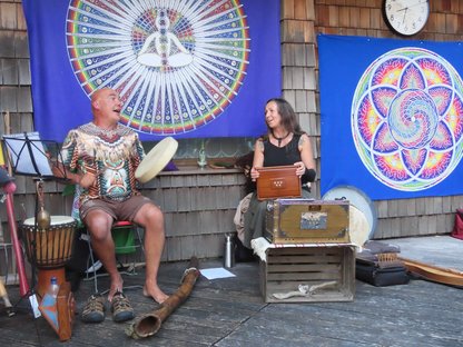 Zwei Musiker, Frau und Mann auf einer Freiluft Holzbühne mit vielen Weltmusik Instrumenten und Mandala Tüchern im Hintergrund 