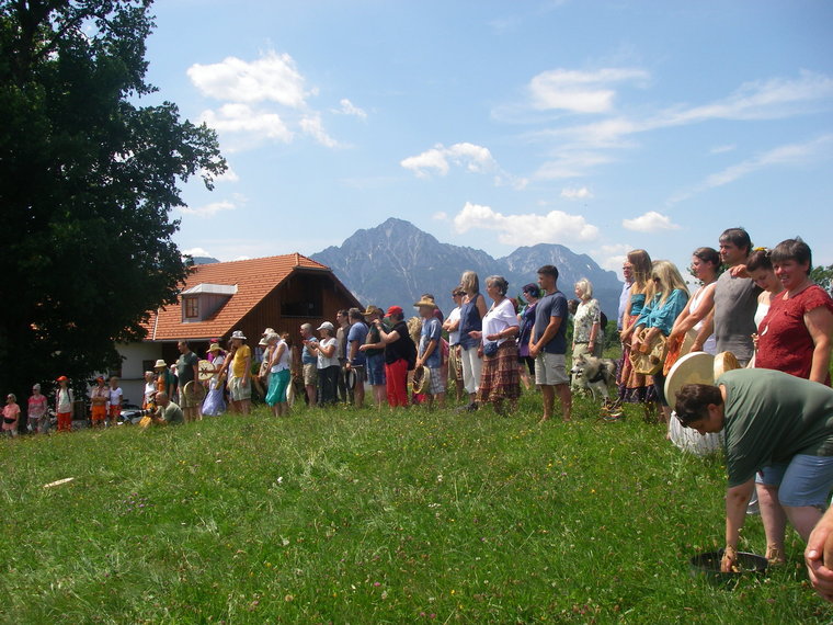 Viele Leute in einer Reihe auf grüner Wiese mit Gasthaus und Berg im Hintergrund