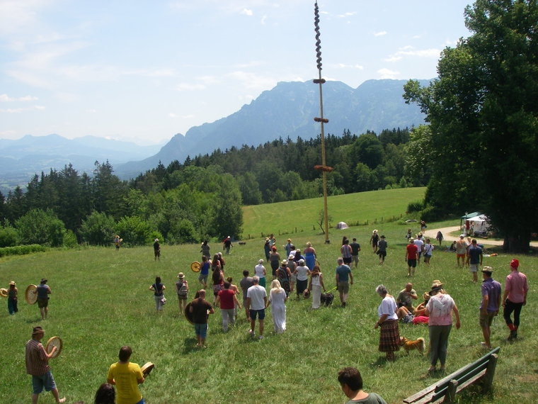 Viele Leute auf sommerlicher Wiese um einen Maibaum und im Hintergrund ein Berg