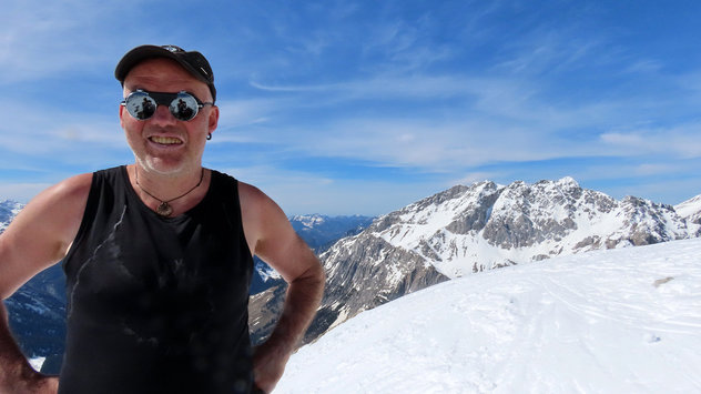 Skibergsteiger in T-Shirt mit Gletscherbrille und schwarzer Kappe auf winterlichen Gipfel im strahlenden Sonnenschein 